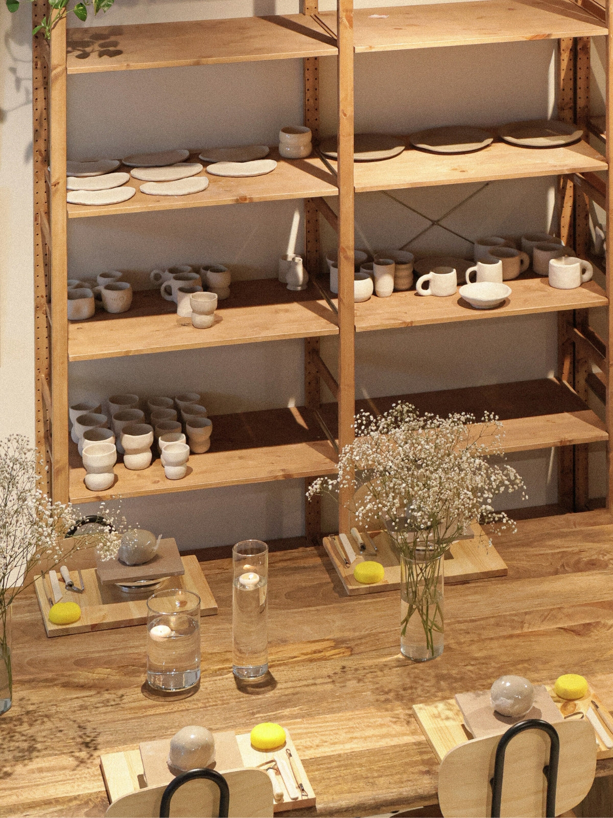 A warm hand-building workshop setup in Dubai, with wooden shelves displaying unfinished ceramic pieces and a table prepared with tools, flowers, and floating candles, from Maison Terraē.